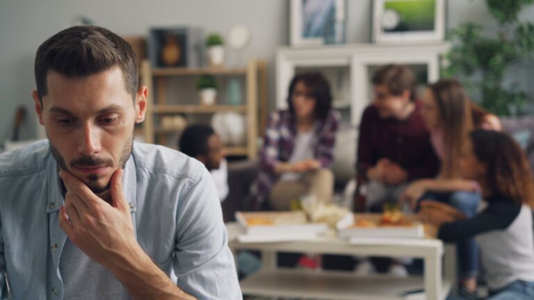 A thoughtful man isolated from friends in a social setting indoors.